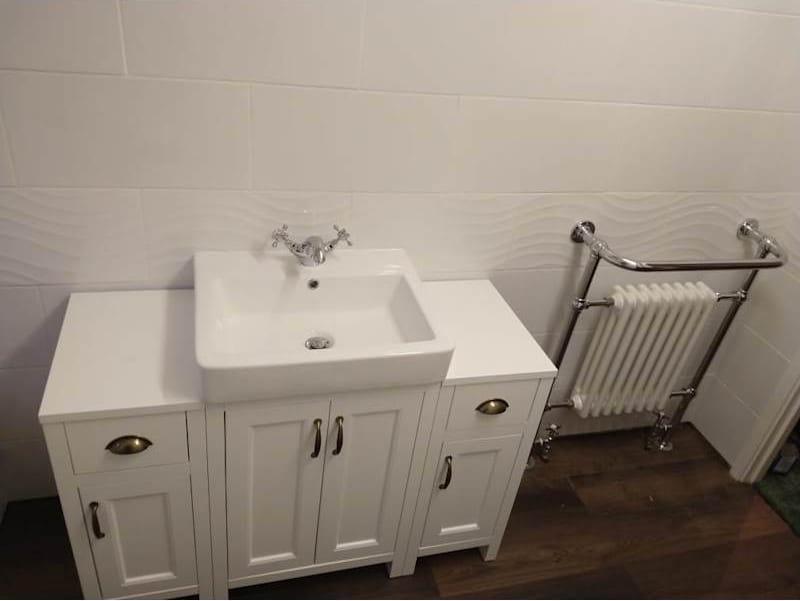 A white vanity unit with a basin set into it, beside a traditional chrome and white towel radiator, against white wave-patterned tiles.