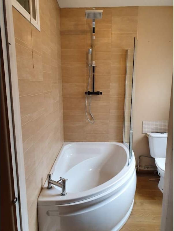 A corner bath with a glass shower screen and a wall-mounted shower, against beige tiling.