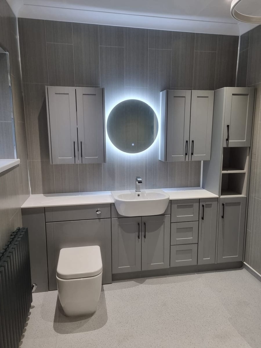 A bathroom fitted by Greg: grey units either side of a round illuminated mirror, a basin set into a white worktop and a back-to-wall toilet.